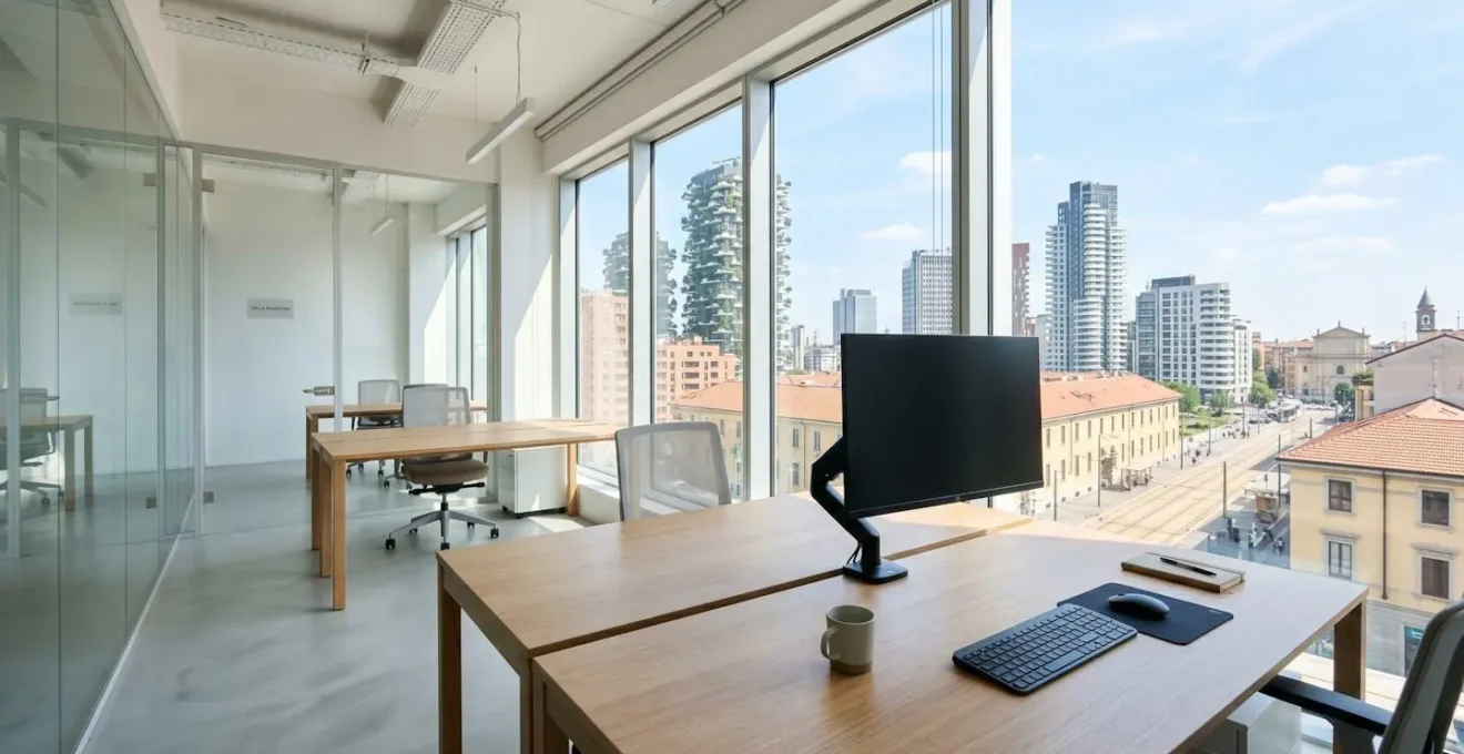Interno di un ufficio moderno italiano vuoto con scrivania pulita, monitor spento e grande finestra che mostra architettura urbana italiana contemporanea, illuminato da luce naturale abbondante